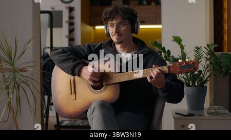 Smiling Caucasian musician guy playing guitar solo at home hipster man in headphones male hobby relaxing music performance song happy artist enjoying Stock Photo