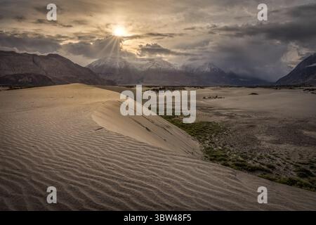 View of stark sand dunes ripple under a dramatic sky, the sun bursting through dark clouds over rugged mountains, Leh, Ladakh, India. Stock Photo