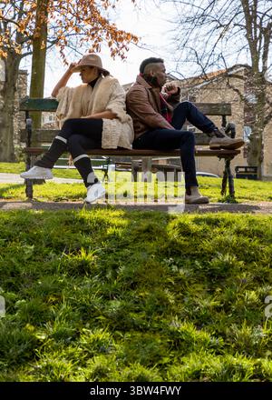 A man and a woman are sitting on a park bench, back to back. The woman is wearing a hat and a fur coat, while the man is wearing a leather jacket and Stock Photo