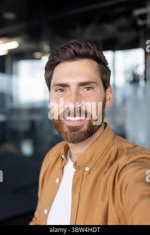 Young successful businessman looking into phone camera, using smartphone app for video call, talking to colleagues and friends while sitting at desk inside office. Stock Photo