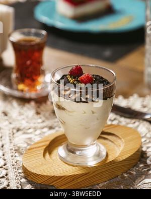 Tiramisu dessert served in an elegant glass, topped with cocoa powder and raspberries, accompanied by a cup of tea on a wooden table with a lace table Stock Photo