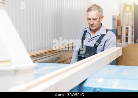 Portrait of an elderly carpenter or carpenter in overalls working with wooden boards in a carpenter's workshop. Sawing wood Stock Photo
