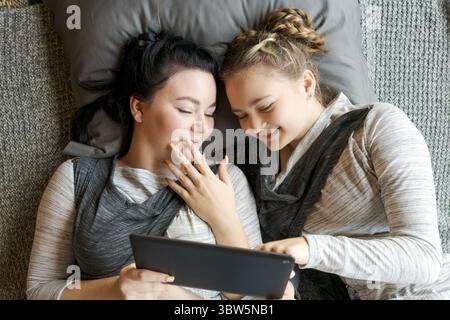 Happy two sisters using tablet while lying on their backs on floor at home laughing cheerfully watching videos or chatting online, wearing grey clothe Stock Photo