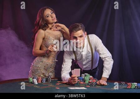 Handsome man and female holding cards, champagne. Playing poker at casino, celebrating their win at table with stacks of chips, cards, on it. Black, s Stock Photo