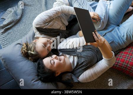 Happy two sisters using tablet while lying on their backs on floor at home laughing cheerfully watching videos or chatting online, wearing grey clothe Stock Photo