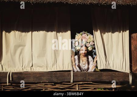Beautiful brunette lady with a long dark hair, in a white ukrainian authentic national costume and a wreath of colorful flowers is smiling and looking Stock Photo