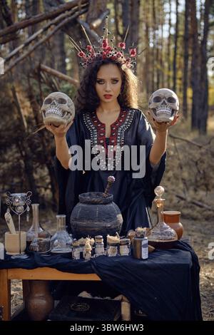 Attractive, wicked, long-haired enchantress in a black, long embroidered dress. There is large red crown in her brown, curly hair. She is holding two Stock Photo