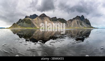 View of majestic, jagged mountains mirroring on the wet, rippled black sand under a brooding, cloudy sky, Hofn, Iceland. Stock Photo