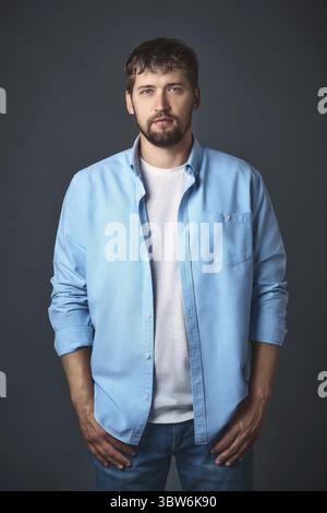 This young confident man with a beard in stylish casual clothes poses in the studio against a gray background. Portrait of a handsome guy of Caucasian Stock Photo