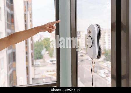Homemaker gesturing toward robotic window cleaner working on tall glass building facade, representing advanced automated maintenance technology in Stock Photo