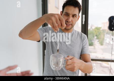 Portrait of happy young man preparing cup of tea while window cleaning robot operates in background, savoring moment of relaxation Stock Photo