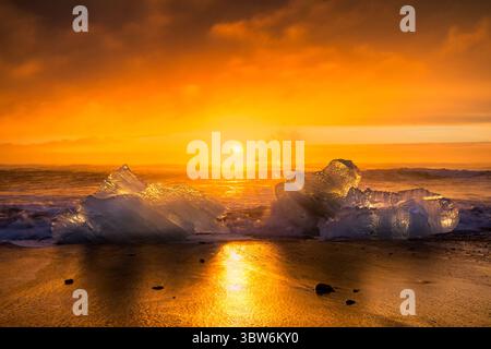 View of radiant sunlight igniting the horizon, casting a golden glow upon the icy sculptures resting on the black sand beach, Vik, Iceland. Stock Photo