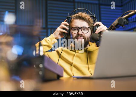 Cheerful young man recording podcast in studio. Confident man with headphones and microphone recording a podcast. Happy guy talking on web radio Stock Photo
