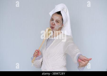 Woman with cosmetic mask on her face, doing anti-aging beauty treatments at home and cheerful sings with a massage brush. Smiling woman in bathrobe an Stock Photo
