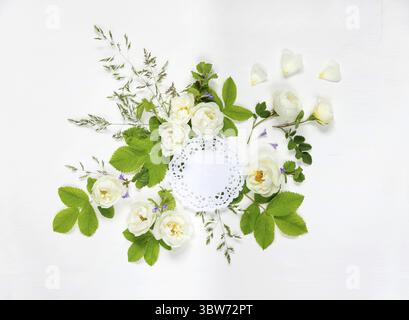 Decorative composition in retro style consisting of round white paper openwork doily and white wild rose flowers with green leaves on white background Stock Photo