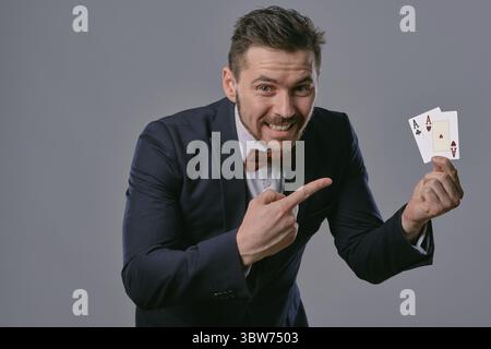 Handsome brunet person in black classic suit and red bow-tie is smiling, showing two playing cards and pointing at them by index finger, posing on gra Stock Photo