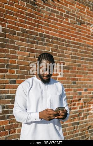 African american man in stylish ethnic dashiki wear viewing photos in social networks on mobile enjoying free time outdoors. Dark-skinned man watching Stock Photo