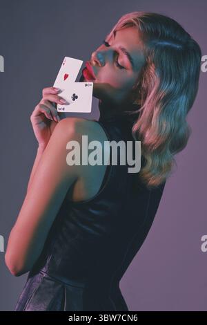 Beautiful blonde lady with bright make-up, in black stylish dress is showing two playing cards and posing sideways against colorful studio background. Stock Photo
