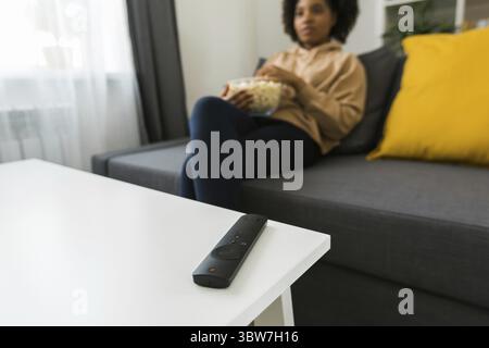 TV remote control on background of African american woman sits on a gray sofa, enjoying a bowl of popcorn while watching a movie or streaming online f Stock Photo