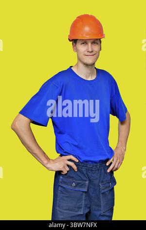Portrait of a young handsome man in a construction helmet and work clothes Isolated on a yellow background. A positive manual laborer Stock Photo