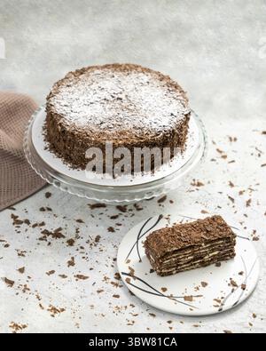 Chocolate cake with chocolate chips sprinkled with powdered sugar standing on a glass stand with a slice on a plate Stock Photo