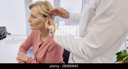 Audiologist putting hearing aid on mature woman's ear at audiology center, close-up, side view. Deafness treatment, hearing solutions Stock Photo