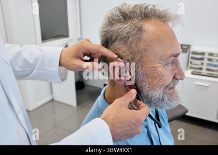 Audiologist carefully adjusts a hearing aid on elderly man using advanced audiological equipment for personalized fitting, side view Stock Photo
