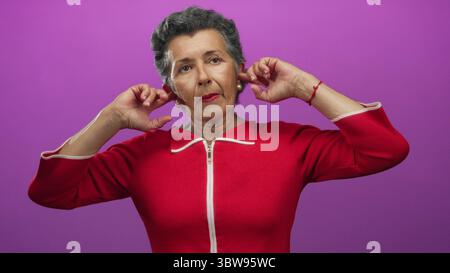 Senior woman with grey hair wearing red blouse standing in front of a vibrant pink background covering her ears with hands in different poses. Stock Photo