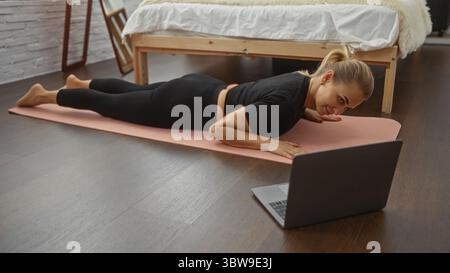 Woman exercising on a yoga mat near bed watching laptop in bedroom home interior featuring blonde adult female doing workout indoors. Stock Photo