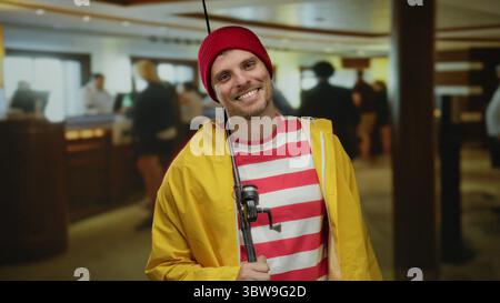 Happy man in striped shirt and yellow jacket with fishing rod stands in a hotel lobby, smiling comfortably, capturing a unique leisure-themed indoor s Stock Photo