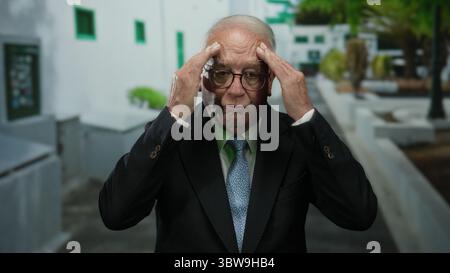 Senior man in business suit stands on an outdoor street, appearing contemplative with hands near face, framed by white buildings and green foliage. Stock Photo
