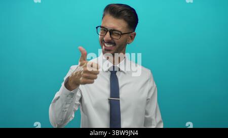 Man in white shirt, tie and glasses giving thumbs up gesture in studio with turquoise backdrop; confidence. Stock Photo