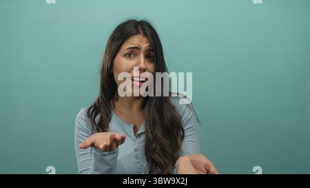 Young hispanic woman beckons open palm for money in studio against green wall, making beckoning gesture; urgent plea. Stock Photo