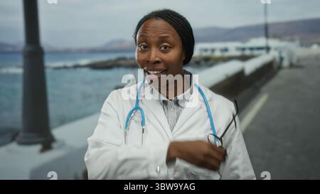 Woman smiling in a white coat with a stethoscope stands on a seaside promenade offering a cheerful professional impression against a blurred ocean bac Stock Photo