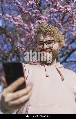 Spring day. Bearded man in pink shirt talking by phone. Spring pink sakura blossom. Handsome young man with smartphone. Fashionable man in trendy glas Stock Photo