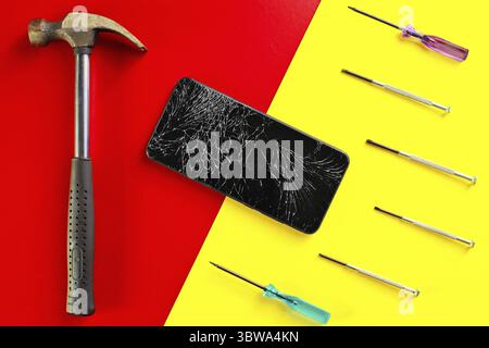 A black touchscreen phone with a cracked display next to screwdrivers and a hammer on a colorful bright background. Top view. Background for repair se Stock Photo