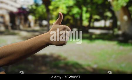 Man giving thumbs-up in a lush outdoor park setting with sunlight filtering through the trees, conveying positivity and relaxation in a natural enviro Stock Photo
