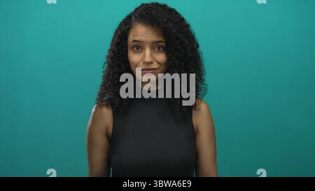 Young hispanic woman with open palms beckons in studio against teal wall with warm smile; friendly invitation. Stock Photo