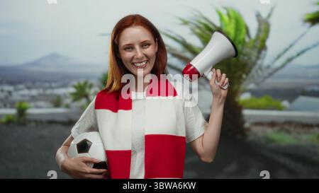 Woman smiling outdoors holding a megaphone and soccer ball, wearing a red and white scarf in a city street, embodying patriotic football enthusiasm. Stock Photo