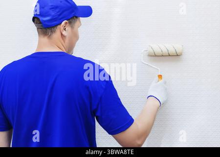 A painter in a cap and gloves paints the wall with a paint roller. Portrait of a young worker in uniform. Professional repair of premises. Mockup Stock Photo