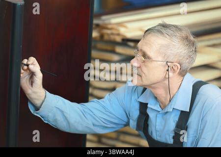 Portrait of an elderly carpenter at work in a carpentry shop. A gray-haired cabinetmaker with glasses is restoring furniture Stock Photo