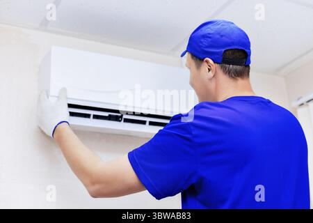 A neat technician in a blue baseball cap and overalls fixes the new air conditioner to the wall in the room. Portrait of a worker from the back. Mocku Stock Photo