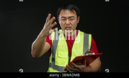 Young asian man wearing safety vest gestures energetically while holding a clipboard against a black background, expressing determination in an isolat Stock Photo