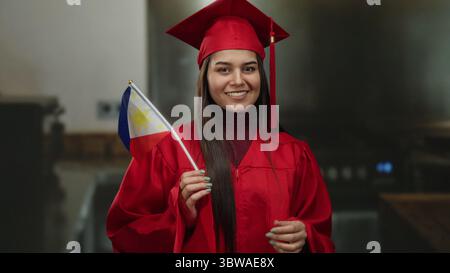 Young woman in a red graduation gown holding a philippines flag smiles proudly indoors, symbolizing academic achievement and cultural pride in a kitch Stock Photo