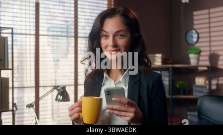 Caucasian woman holding cup coffee using mobile phone smartphone drinking smile female girl businesswoman business office browsing online scrolling Stock Photo