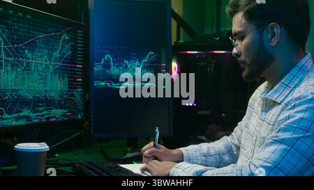 Indian Arabian man writing notes notebook working with pc computer trader programmer coder hacker male complete finish work online businessman night Stock Photo