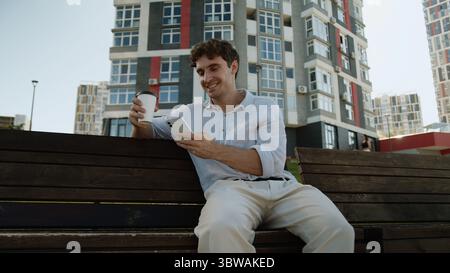 Caucasian young man on bench using mobile phone smartphone app happy male holding cup of coffee drink at city relaxed guy smiling student break Stock Photo