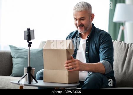Happy senior man blogger broadcasting while opening delivery box Stock Photo