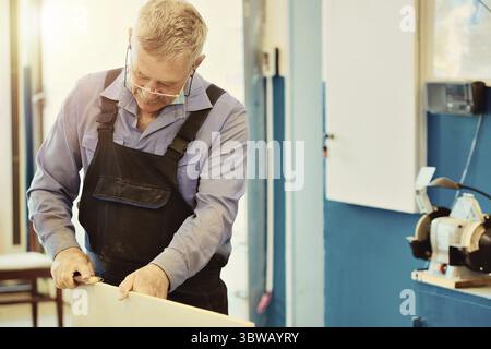Portrait of an elderly carpenter or carpenter in overalls working with wooden boards in a carpenter's workshop. Sawing wood Stock Photo