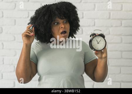 Surprised african american woman holding alarm clock amazed with open mouth and surprise happy face at home, on brick background. Being late and time Stock Photo